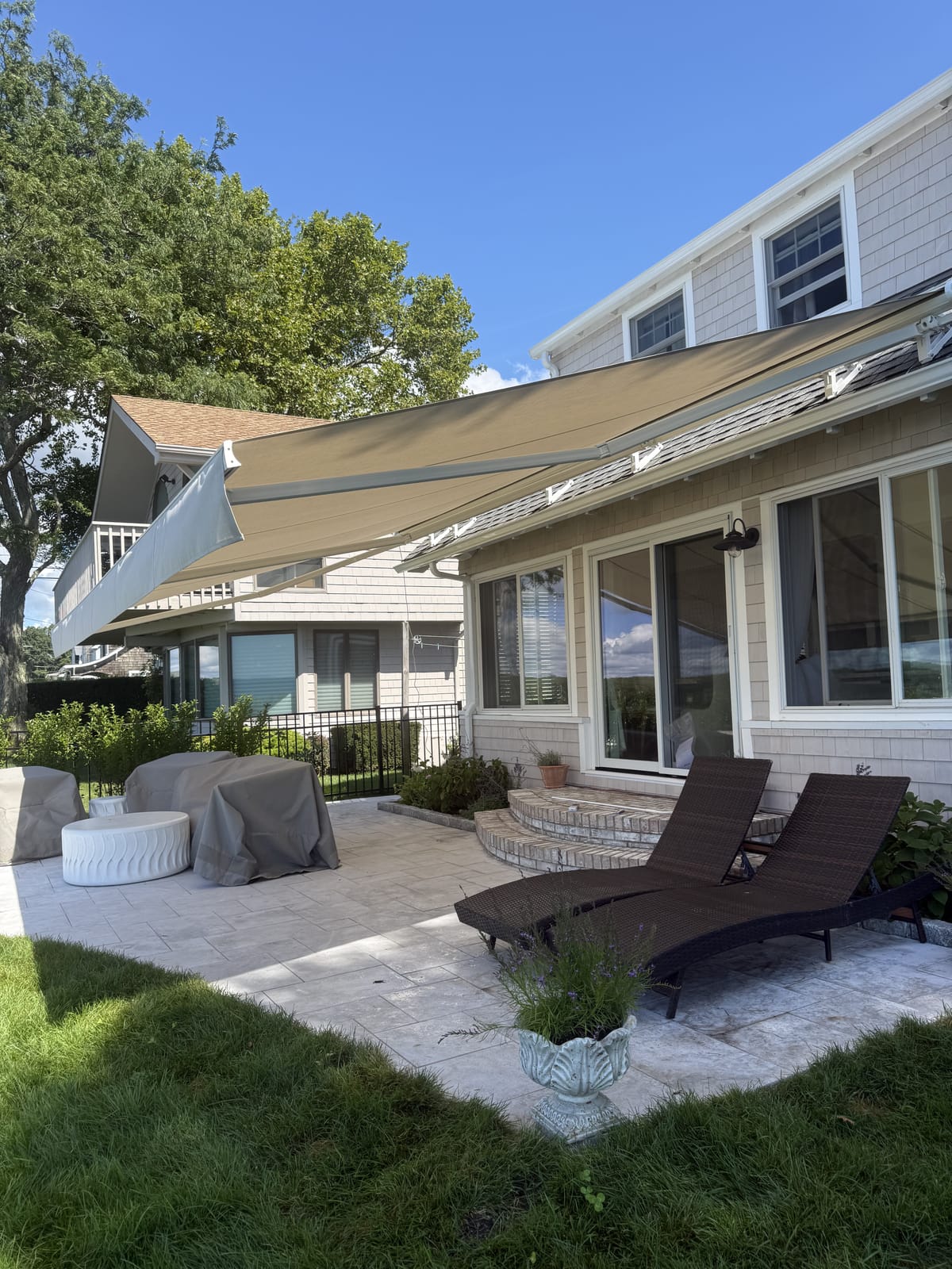 Sand-colored retractable awning extended over a stone patio at a shingle-style coastal home