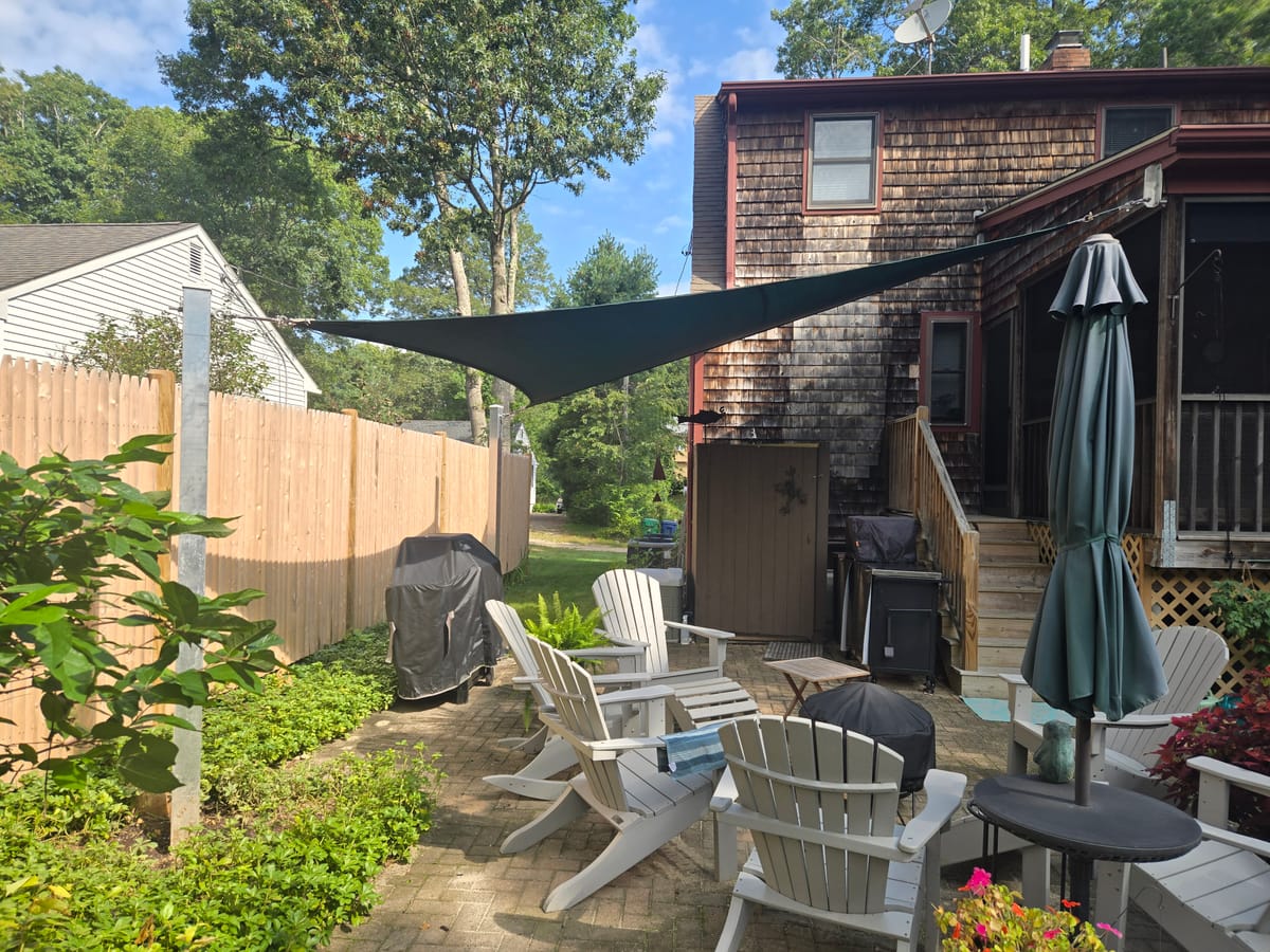 Green triangular shade sail over a backyard paver patio with Adirondack chairs, anchored between a steel post and a cedar-shingled home