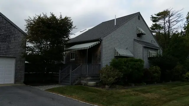 Striped window awnings shading the porch entry and gable windows of a gray shingle cottage
