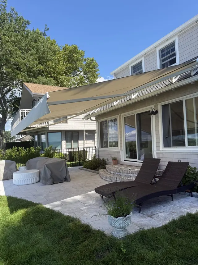 Sand-colored retractable awning extended over a stone patio at a shingle-style coastal home