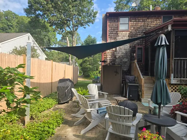 Green triangular shade sail over a backyard paver patio with Adirondack chairs, anchored between a steel post and a cedar-shingled home