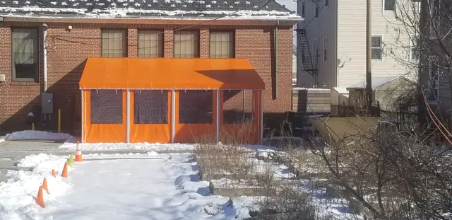 Enclosed orange canopy with clear vinyl curtains outside a brick building, mid-winter